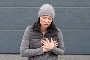 Woman standing outdoors in a grey winter outfit clutching her chest with both hands and grimacing in pain in a concept of a heart attack or cardiac problem