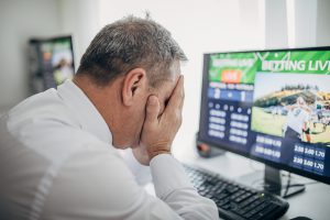 A man holding his face in his hands in distress while using a computer for online gambling.