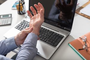 A man holding his wrist in front of his laptop due to pain from carpal tunnel syndrome.