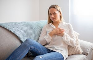 A woman sitting on a couch holding her chest due to a heart murmur.