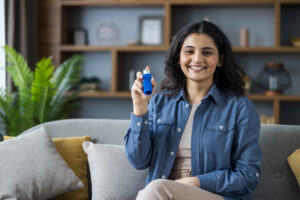 Portrait of a young smiling Muslim woman sitting on the sofa at home in a shirt, holding an inhaler for breathing, and showing a bottle of medicine to the camera.