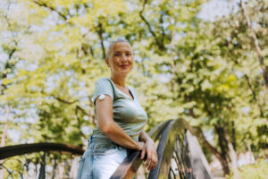 Woman with a visible diabetic sensor on her arm poses for the camera while leaning on a bridge in a public park, enjoying a bright and sunny day.