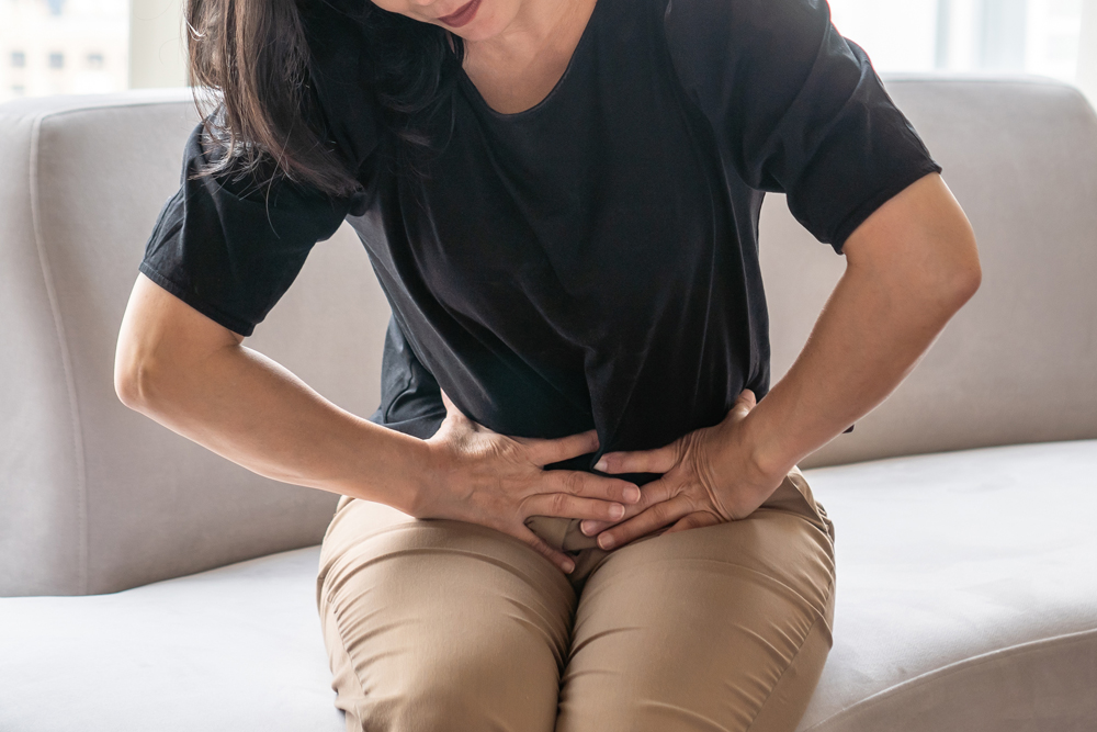 Abdominal pain in woman with stomachache illness from menstruation cramps, stomach cancer, irritable bowel syndrome, pelvic discomfort, Indigestion, Diarrhea or GERD (gastro-esophageal reflux disease) A woman bent over and holding her lower abdomen while sitting on a couch.