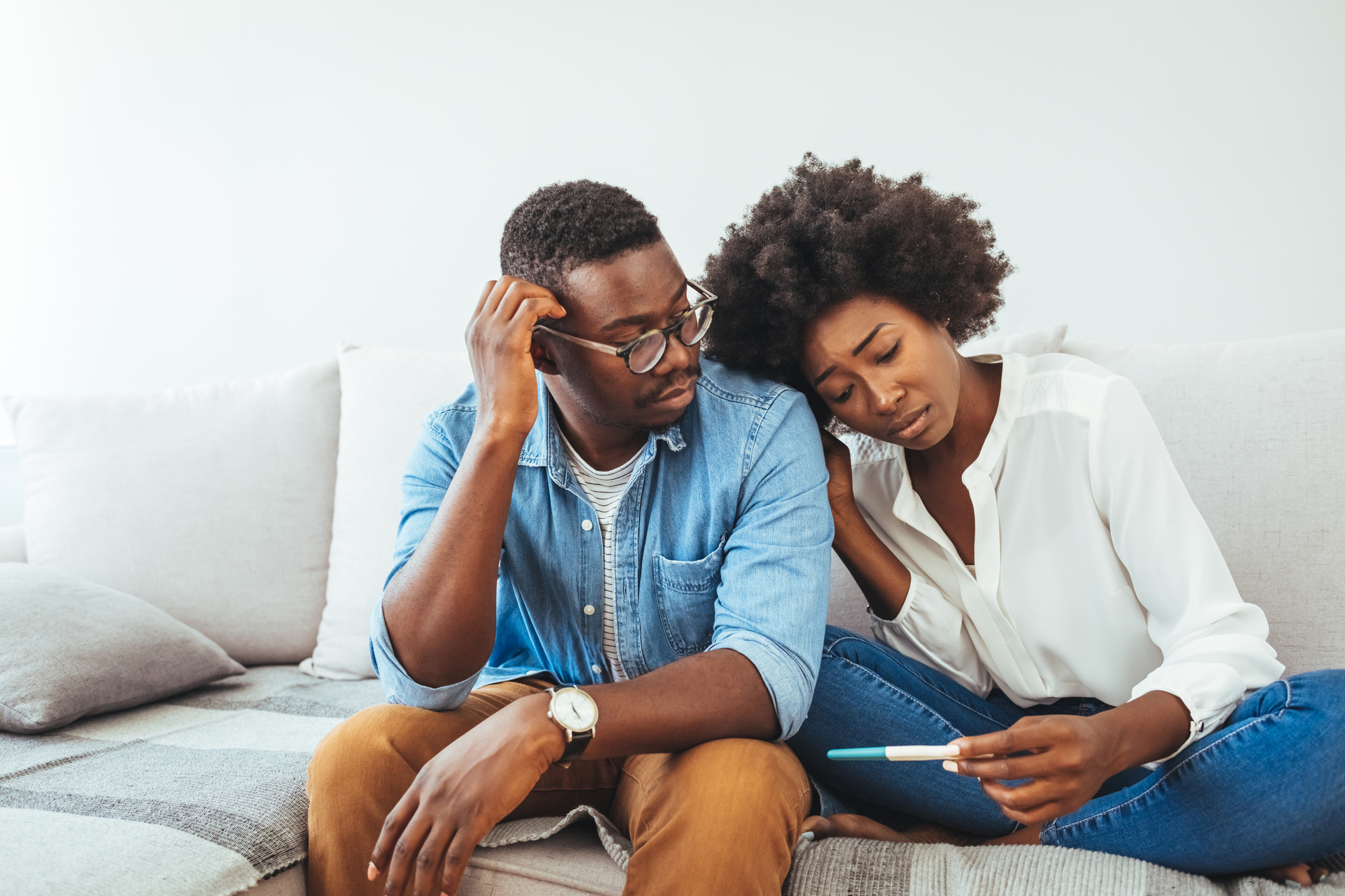 A sad young woman holds a pregnancy test in her hand. A couple sitting on a couch looking down sadly at a pregnancy test result.