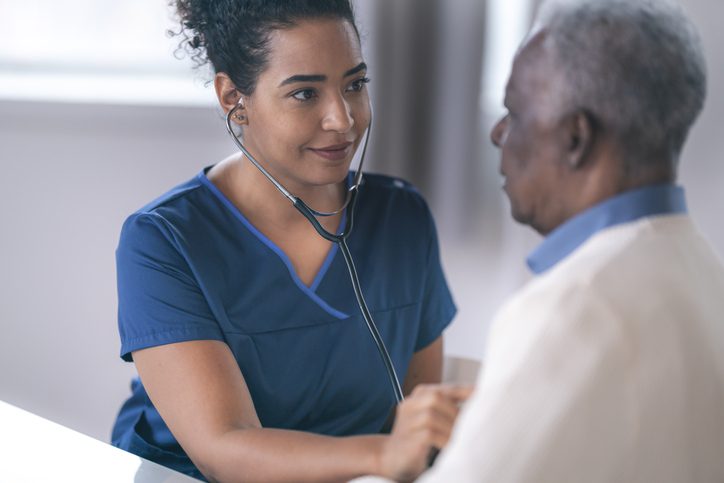 A female black doctor is performing a routine medical exam. The patient is a senior adult man. The two individuals are seated facing each other in an examination room. The doctor is using a stethoscope to check the patient's heart and lungs. The close up image is focused on the smiling doctor.