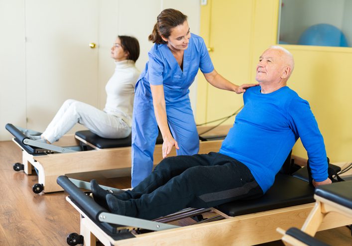 Young woman physiatrist helping aged man doing exercises on reformer as part of injury rehabilitation. Wellness concept for elderly